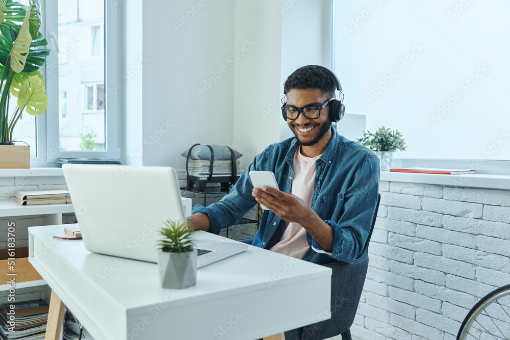 Happy young African man in headphones using smart phone while working in office