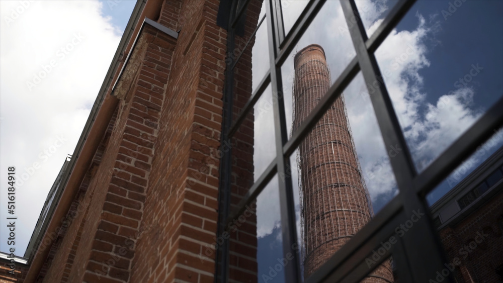 Old chimney of Moscow Arma business quarter reflected in a window. Action. Bottom view of a red brick wall of an old gas factory with a high chimney on blue cloudy sky background.