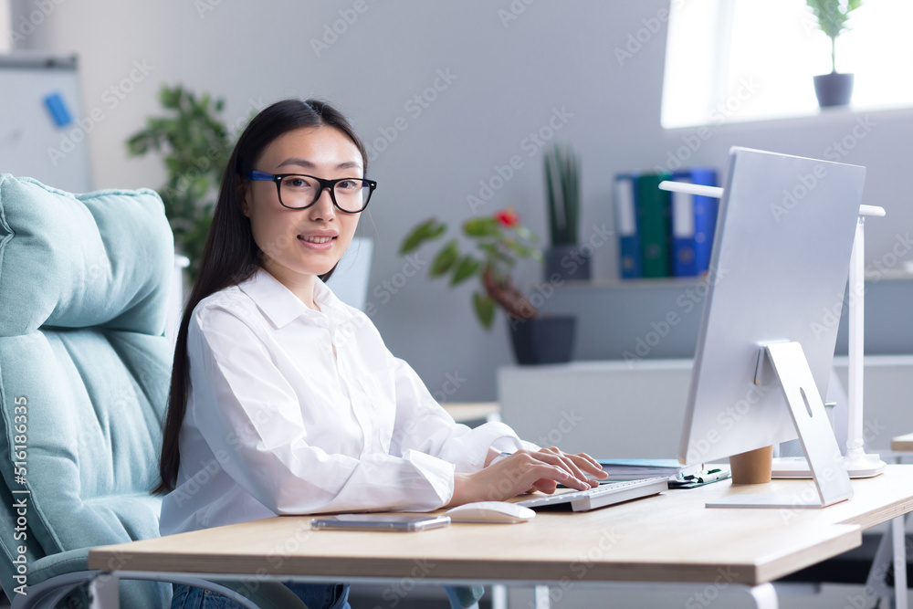 Portrait of a young Asian woman in the office. Businesswoman, director, freelancer, manager, sitting in a white shirt and glasses at the desk, working at the computer. He looks at the camera, smiles. Portrait of a young Asian woman in the office. Businesswoman, director, freelancer, manager, sitting in a white shirt and glasses at the desk, working at the computer. He looks at the camera, smiles.