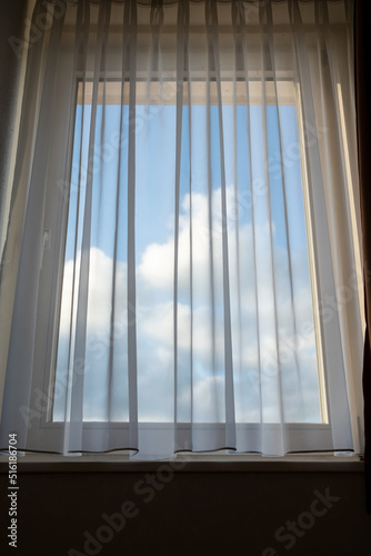 Closed window with a transparent curtain through which you can see the blue sky and clouds. Close-up.