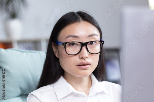 Close-up photo. Portrait of a young Asian woman in the office. Businesswoman, director, freelancer, manager, sitting in a glasses at the desk, working at the computer. He looks at the camera.