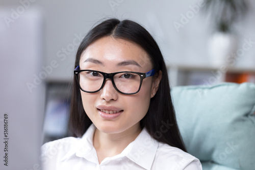 Close-up photo. Portrait of a young beautiful Asian woman in the office. Businesswoman, director, freelancer, manager, sitting in a white shirt and glasses at the desk, working at the computer.