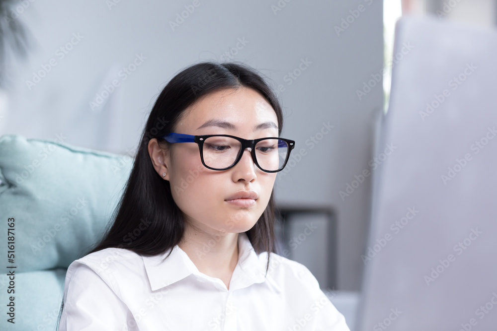 Close-up photo. Portrait of a young beautiful Asian woman in the office. Businesswoman, director, freelancer, manager, sitting in a white shirt and glasses at the desk, working at the computer. Close-up photo. Portrait of a young beautiful Asian woman in the office. Businesswoman, director, freelancer, manager, sitting in a white shirt and glasses at the desk, working at the computer.