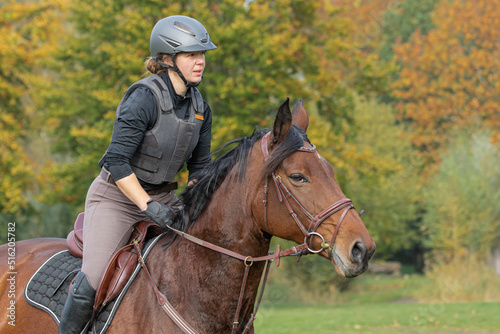 junge Frau im leichten Sitz mit Sicherheitsweste und Reithelm galoppiert auf braunem Pferd bei Ausritt im Herbst