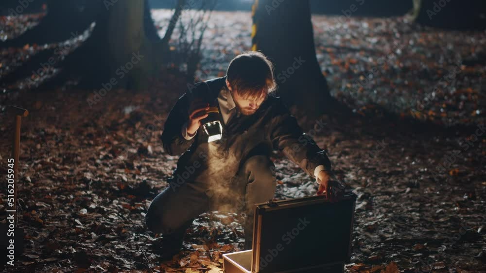 Young smart bearded man with flashlight in hand opening black briefcase in forest at night. Secrets, creepy story. Detective looking for evidence of crime