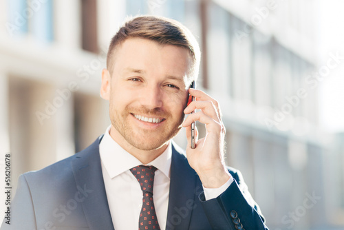 Portrait of delightful European businessman, wears elegant suit, talks on smart phone, deals with business partner, decide time of meeting, has positive expression. People, career, technology concept photo