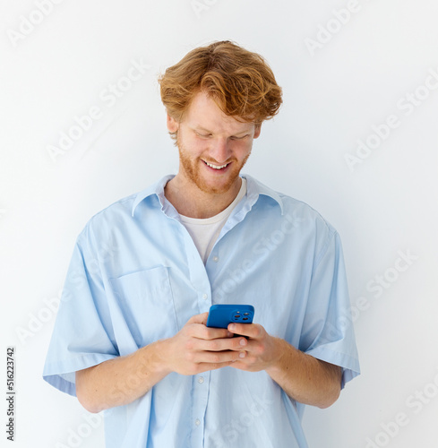 Man using mobile phone. Young redhead man holding smartphone standing on a white background; friendly smiling; typing a message; online chatting