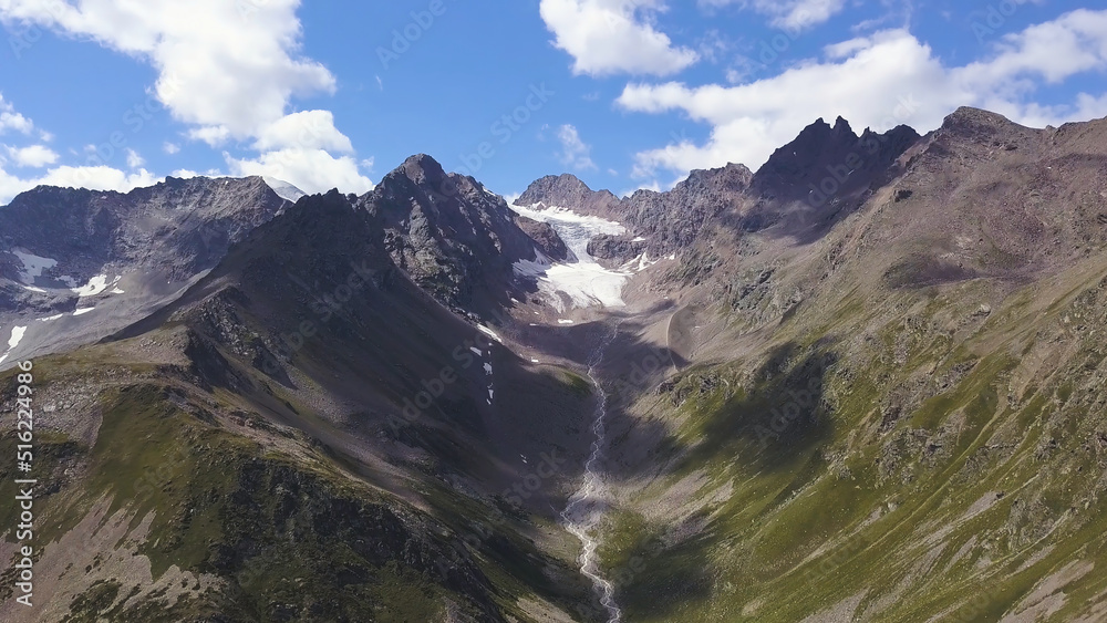 Awesome aerial view to big snowy rocky mountains under blue sky. Clip. Wonderful scenery with giant mountains with green slopes and white peaks and the gorge between two ranges.