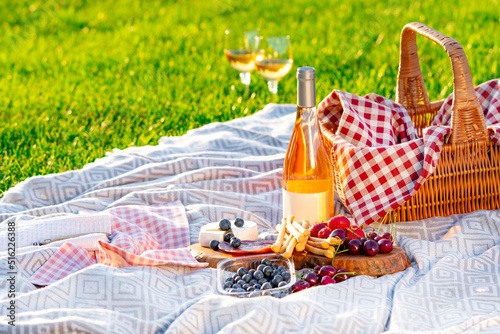 Picnic setting in a meadow. Picnic basket, wine, berries, jamon, cheese and bread are on the blanket.
