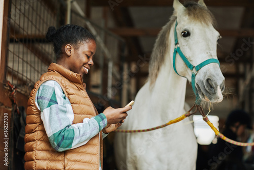 Side view portrait of smiling African American woman using smartphone in horse stables with white stallion in background, copy space