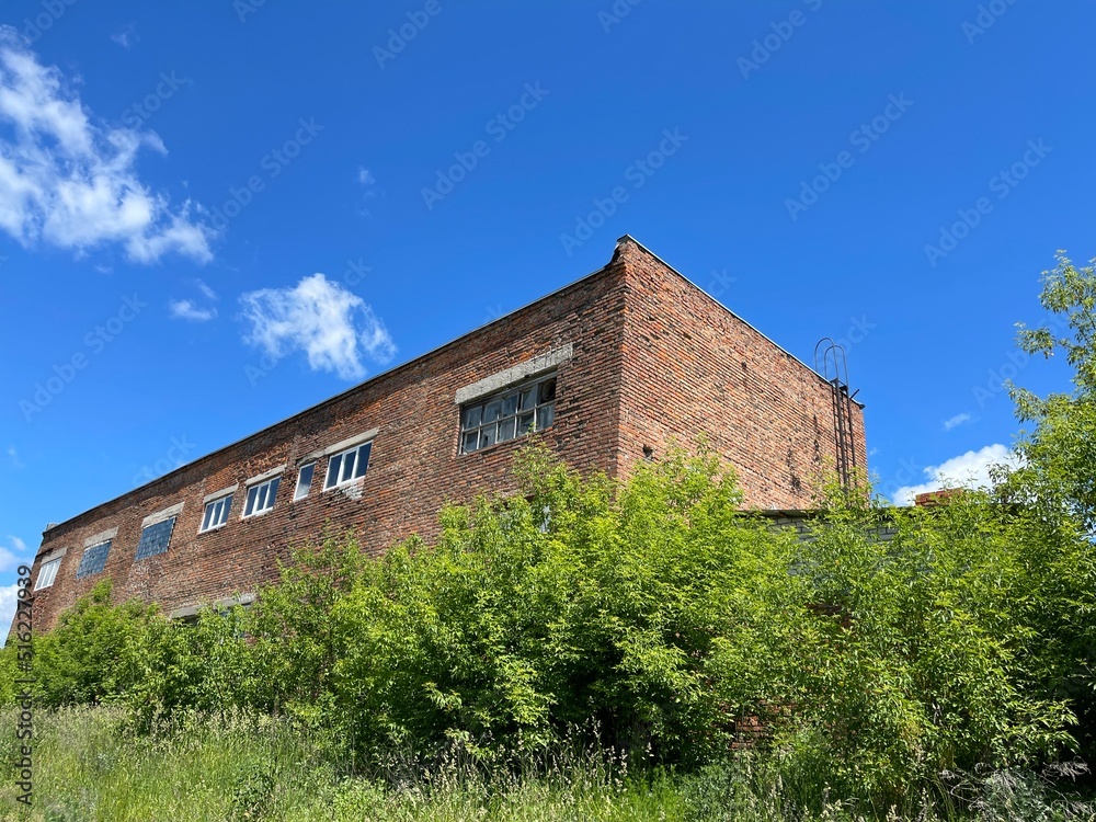 Old brick industrial building in sunny weather. Building exterior of shabby unkempt building in countryside on background of blue sky with white clouds. Old brick industrial building in sunny weather. Building exterior of shabby unkempt building in countryside on background of blue sky with white clouds.