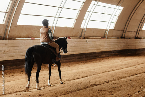 Wide angle view at young woman riding horse in indoor arena lit by soft light, copy space
