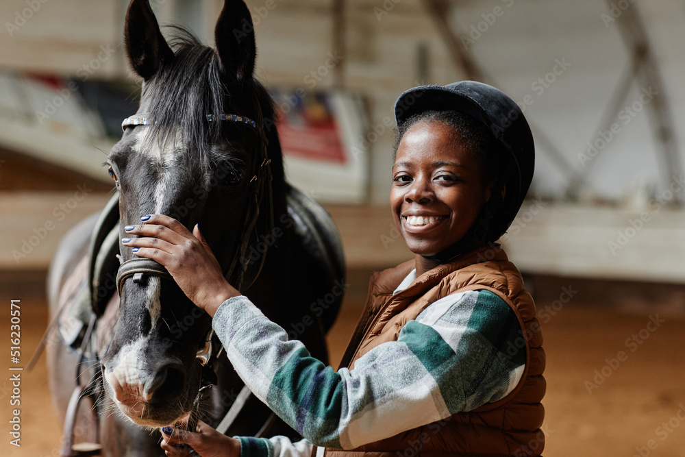 Waist up portrait of smiling black woman posing with horse in indoor riding arena and looking at camera, copy space
