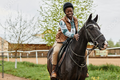 Portrait of smiling black woman riding horse in outdoor country ranch and stroking gently, copy space © Seventyfour