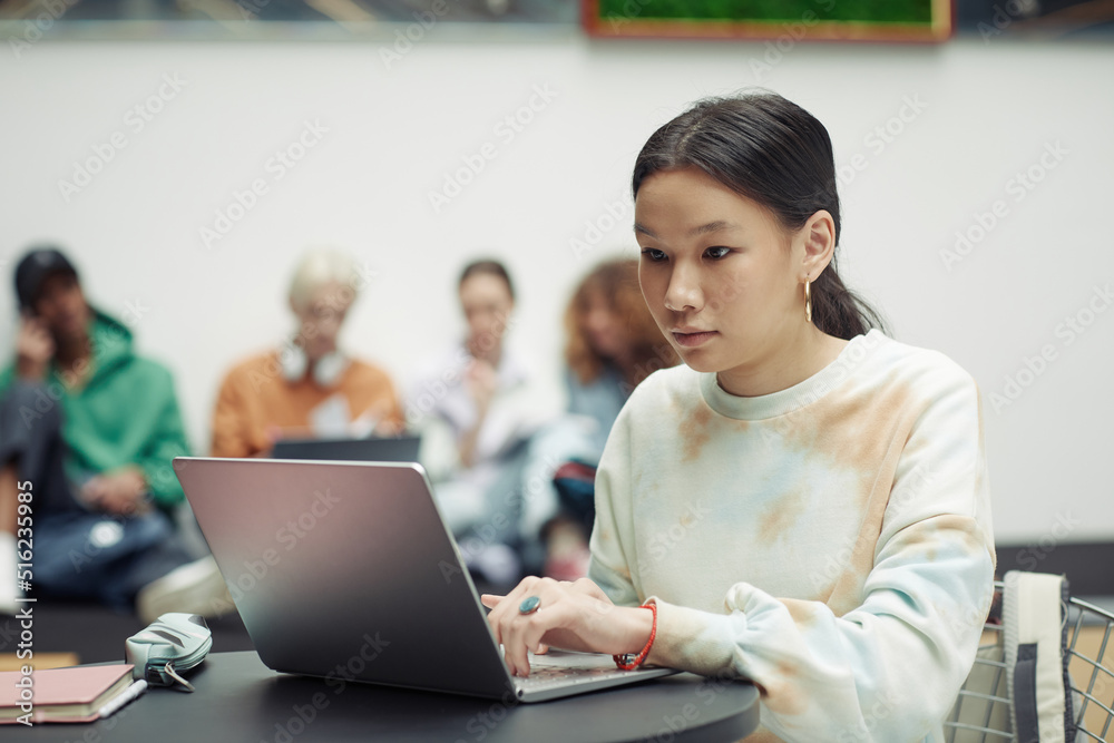 Asian teenage girl in white sweatshirt looking at laptop screen and touching keypad while working over school assignment Asian teenage girl in white sweatshirt looking at laptop screen and touching keypad while working over school assignment