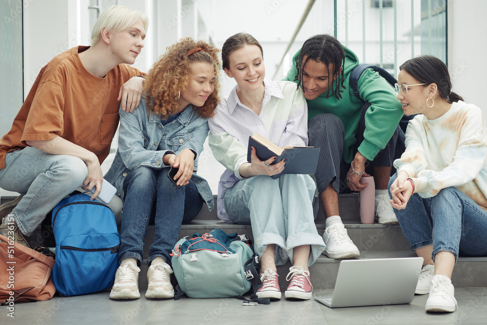 Large group of teenagers in casualwear reading open book held by smiling girl and discussing paragraph before lesson Large group of teenagers in casualwear reading open book held by smiling girl and discussing paragraph before lesson