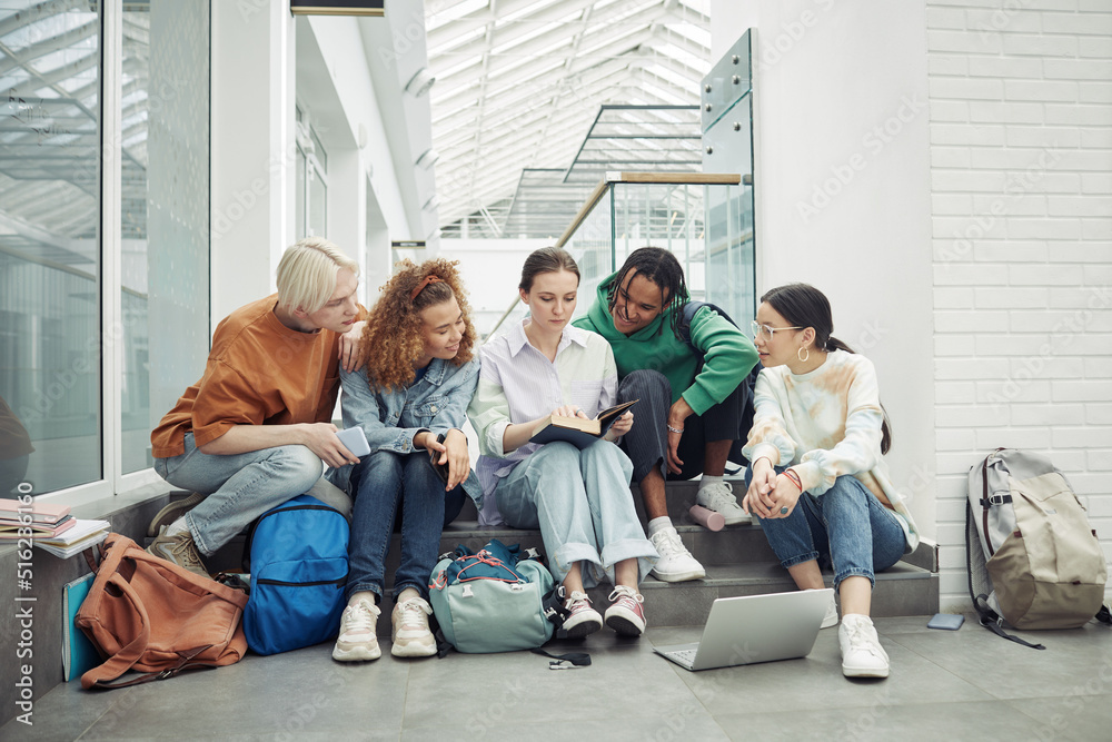 Group of diligent teenage students looking through paragraph in textbook held by girl in jeans, gumshoes and white shirt Group of diligent teenage students looking through paragraph in textbook held by girl in jeans, gumshoes and white shirt