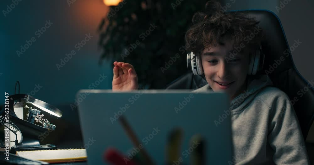 The boy has remote school activities in room, sitting comfortably in desk chair with wireless headphones in front of laptop. He waves hand in greeting to teacher and classmates.