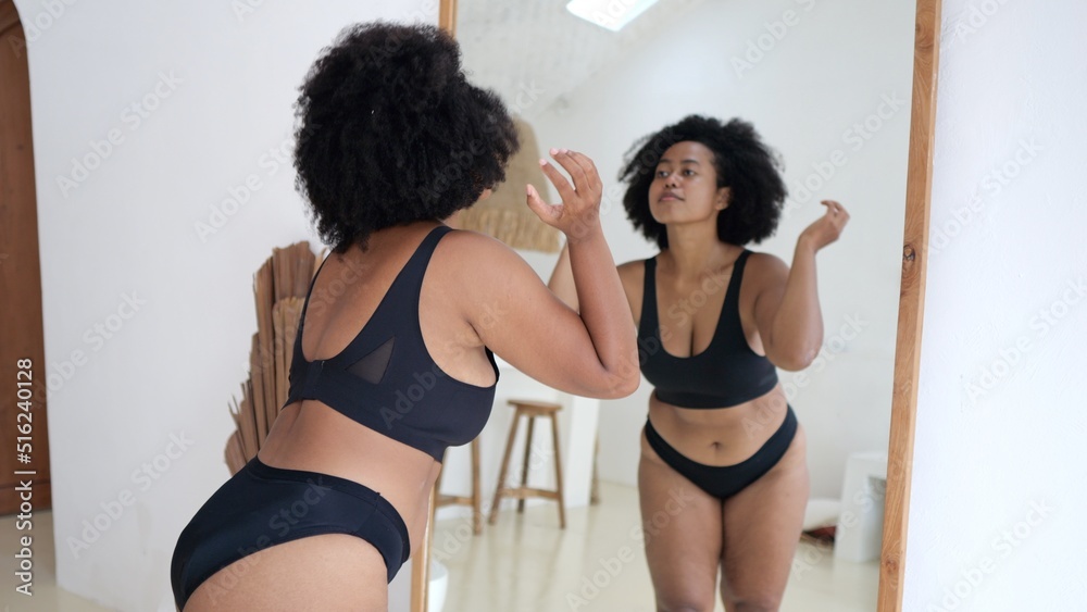 Multicultural plus size woman in lingerie looking at her body in the  mirror. Black woman struggles