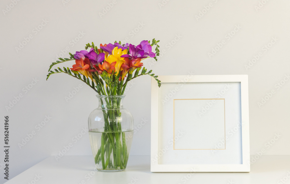 Close up of colourful freesia flowers in glass vase with blank square picture frame against white wall (selective focus) Close up of colourful freesia flowers in glass vase with blank square picture frame against white wall (selective focus)
