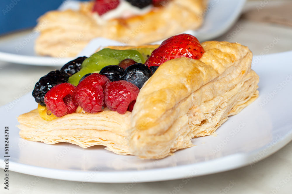 A square fruit filled French puff pastry on a white square plate. The cream and custard filled dessert have strawberries, kiwi, and blackberries on top with a sugar glaze. The pastry is flaky. A square fruit filled French puff pastry on a white square plate. The cream and custard filled dessert have strawberries, kiwi, and blackberries on top with a sugar glaze. The pastry is flaky.