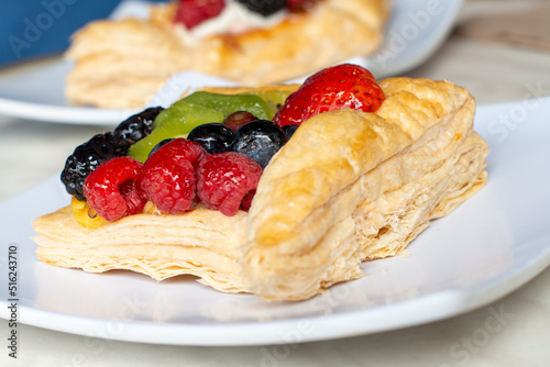 A square fruit filled French puff pastry on a white square plate. The cream and custard filled dessert have strawberries, kiwi, and blackberries on top with a sugar glaze. The pastry is flaky.	