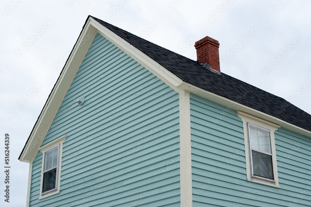 An upward view of a vintage blue wooden building with thick white trim, a small double hung window, with four panes of glass, and a frame shingled roof. The roof has white trim and black shingles.