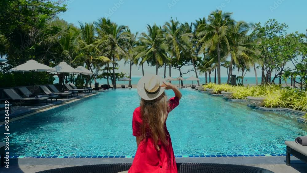 Summer travel, holidays, vacation concept. Back view of young woman in red dress wearing straw hat resting by amazing blue swimming pool in luxury hotel or resort with palm trees, sea background.