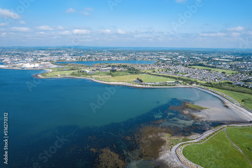 Aerial view on Galway city and Ballyloughane Strand. Warm sunny day. Blue sky and water of the ocean. High tide. Residential area and sport ground. Popular rest area with foot path for walk.