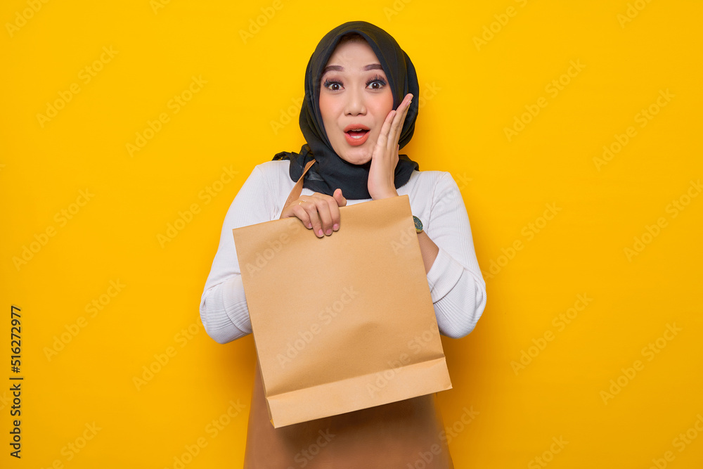 Excited young Asian woman barista in white t-shirt apron work in coffee shop hold blank paper takeaway bag mock up isolated on yellow background. Business startup Excited young Asian woman barista in white t-shirt apron work in coffee shop hold blank paper takeaway bag mock up isolated on yellow background. Business startup