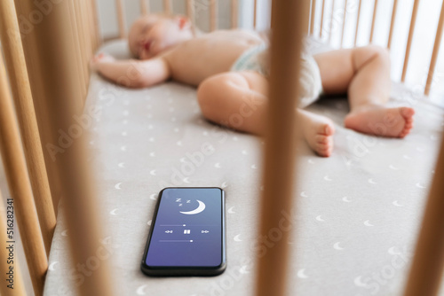 Fotografía Selective focus of little boy in crib falling asleep with the help of application for baby sleep on phone lying next to him while mother cooking dinner at kitchen