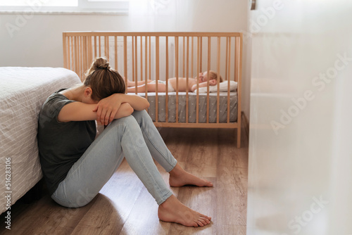 Young mother suffering postnatal depression sitting on floor next to baby napping in bed, resting head on knees, crying, feeling desperate in need of professional psychological support photo