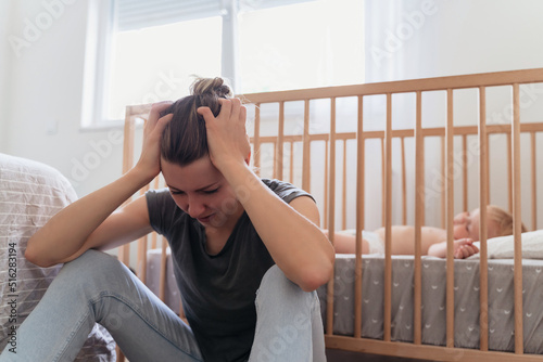 Young mother holding head in hands with desperation, feeling tired and exhausted, crying sitting next to baby sleeping in bed, suffering postnatal depression in need of psychological treatment