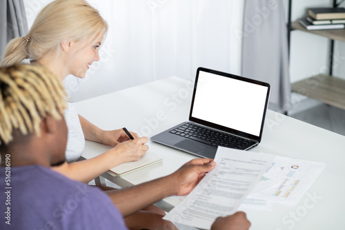 Virtual meeting. Working couple. Digital mockup. Smiling woman making notes while black man checking documents sitting desk with laptop blank screen in light room interior.