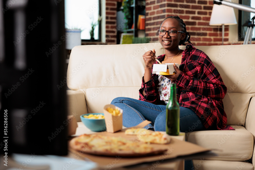African american woman eating noodles from takeout delivery package and relaxing on couch with movie on tv. Using choppsticks to eat takeaway fast food meal and drinking beer from bottle. African american woman eating noodles from takeout delivery package and relaxing on couch with movie on tv. Using choppsticks to eat takeaway fast food meal and drinking beer from bottle.