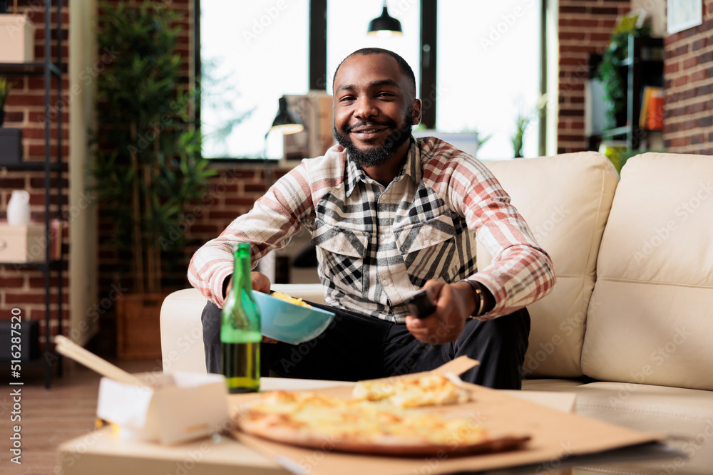 Young adult switching tv channel with remote control and eating chips, finding movie to watch on television at home. Enjoying snacks and beer bottles with takeaway delivery food on sofa. Young adult switching tv channel with remote control and eating chips, finding movie to watch on television at home. Enjoying snacks and beer bottles with takeaway delivery food on sofa.