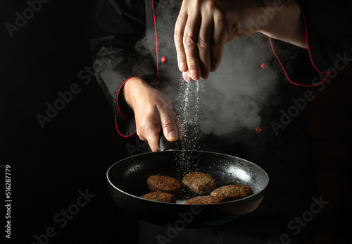 Fototapeta Naklejka Na Ścianę i Meble -  The chef adds salt to cutlets that are fried in a pan. Cooking meat patties for a burger. Menu for a restaurant or hotel on a black background.