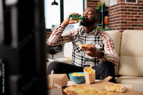 Young adult drinking alcoholic beer from bottle and eating takeaway meal from fast food delivery package, watching movie on tv. Enjoying drink and takeout meal in front of television.