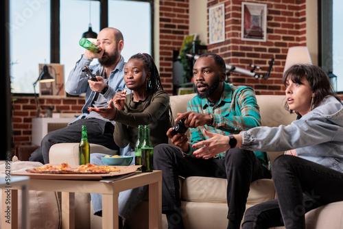 Group of diverse friends playing video games together, having fun at home gathering with beer and food. Enjoying gaming competition on television console, people cheering and drinking.