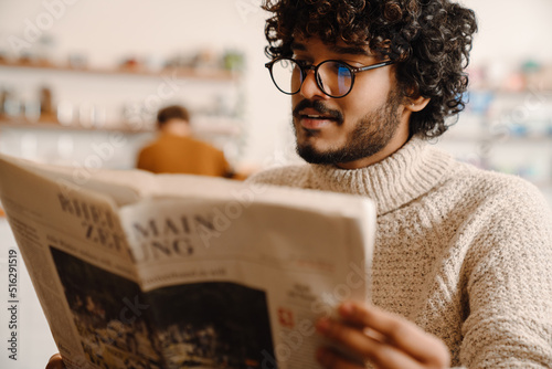 Young handsome indian curly man in glasses reading newspapper photo