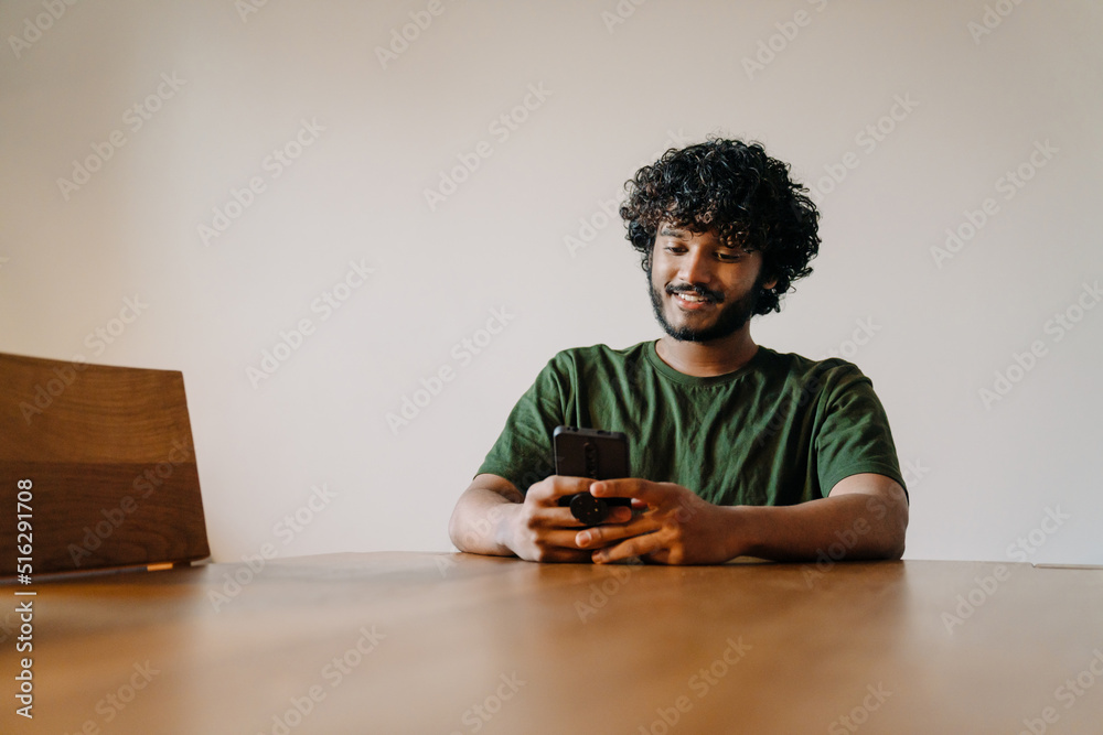Young indian handsome curly man sitting by table with phone
