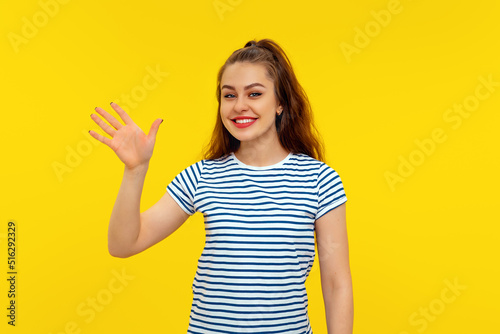 Friendly cheerful brunette girl smiling, saying hi and waving hand to greet person, make hello gesture, welcome someone, standing in white-blue striped t shirt over yellow background