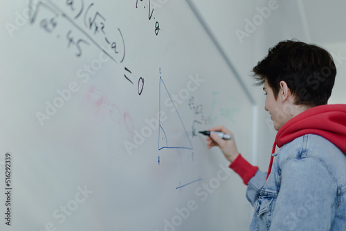 Back view of high school student solving math problem on whiteboard in classroom.