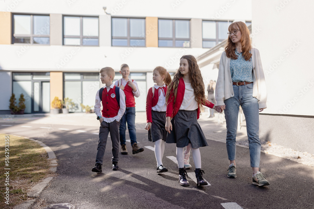 Fototapeta premium Happy schoolkids in uniforms with teacher walking at a schoolyard.