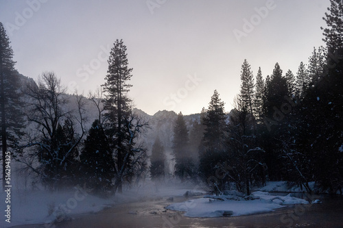 Yosemite valley is enshrouded in a thin layer of mist hanging over the merced river, providing an eerie atmosphere around sunset.
