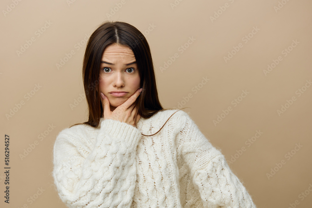 a disgruntled woman is standing sideways on a light brown background in a white sweater, looking forward with condemnation, raising her finger to her chin a disgruntled woman is standing sideways on a light brown background in a white sweater, looking forward with condemnation, raising her finger to her chin