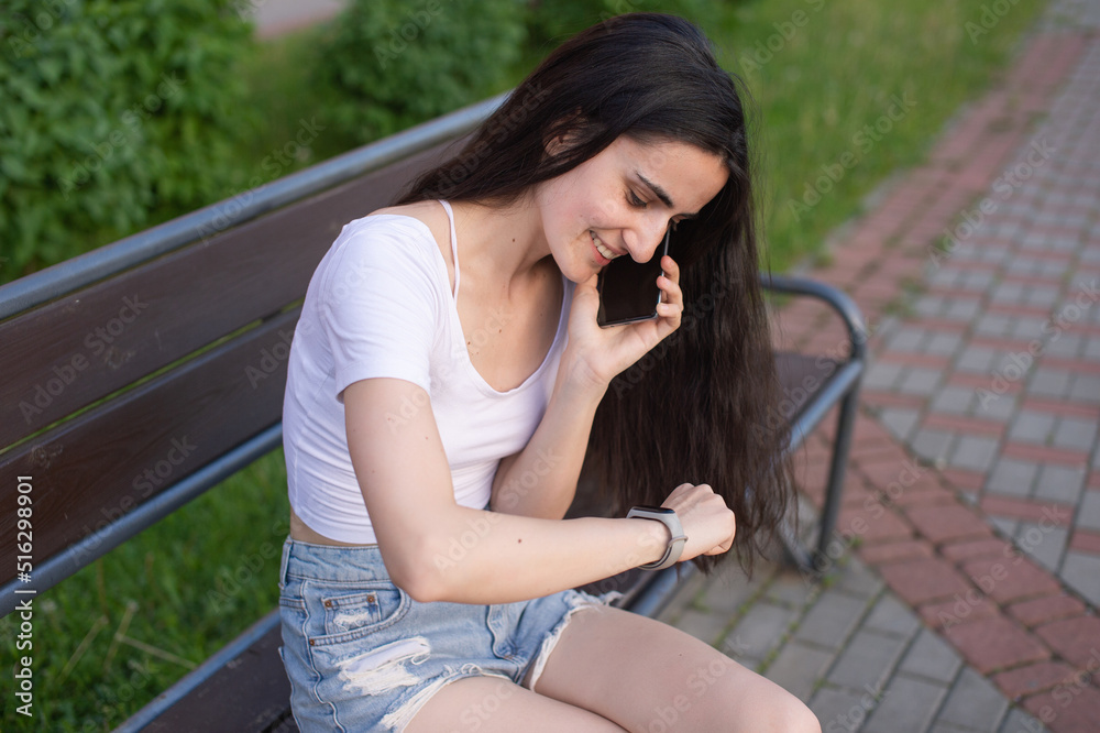Naklejka premium The girl is sitting on a bench, laughing and looking at the phone, there is a laptop nearby