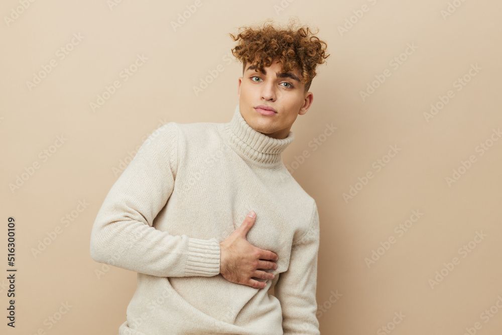 portrait of a handsome man with curly hair, standing on a beige background in a light turtleneck, holding his hand on his body, looking pleasantly into the camera. portrait of a handsome man with curly hair, standing on a beige background in a light turtleneck, holding his hand on his body, looking pleasantly into the camera.