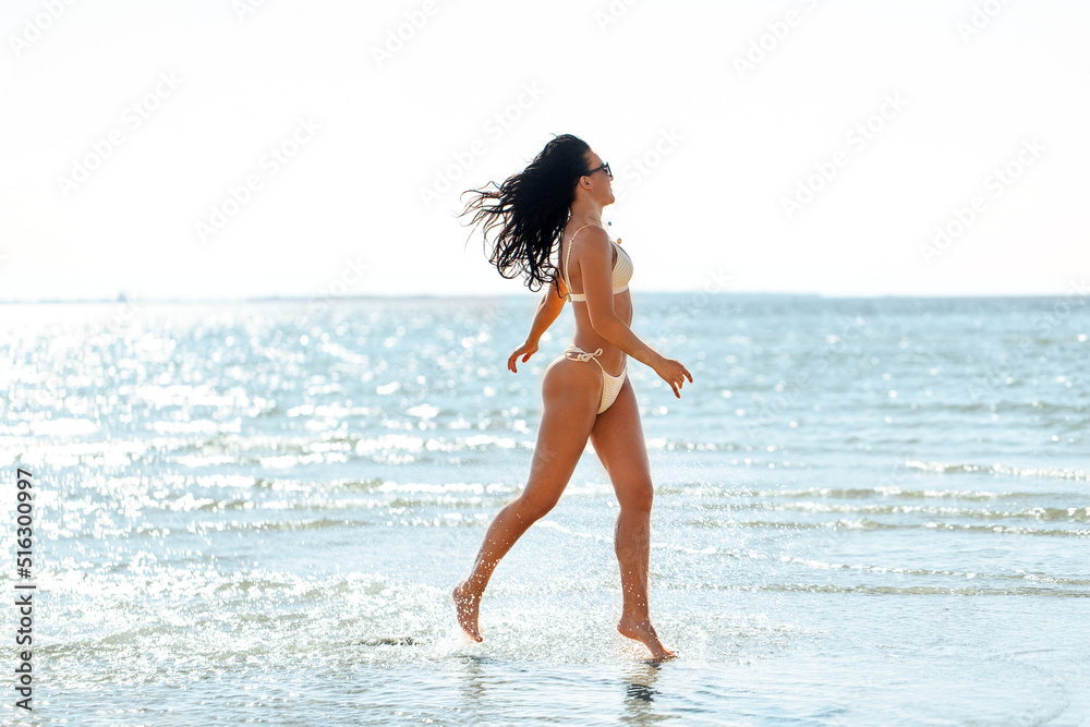 Naklejka premium people, summer and swimwear concept - young woman in bikini swimsuit running in shallow water on beach