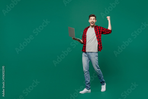 Full body young happy smiling caucasian man he 20s wearing red shirt grey t-shirt hold use work on laptop pc computer isolated on plain dark green background studio portrait. People lifestyle concept © ViDi Studio